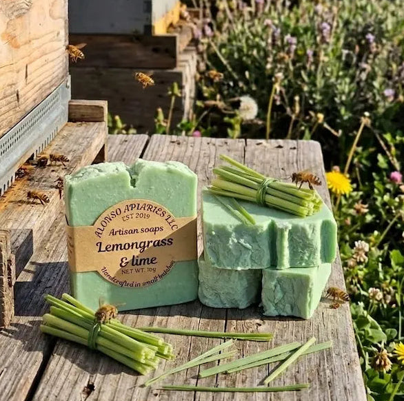 Two green soap bars with lemongrass and lime scent on a wooden surface with flowers and bees in the background.